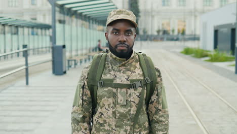 Portrait-of-serious-African-American-handsome-young-male-soldier-in-cap-with-backpack-standing-at-street.-Man-militarian-looking-at-camera-at-train-station.-Bus-stop.-Military-uniform.-Zooming-in.